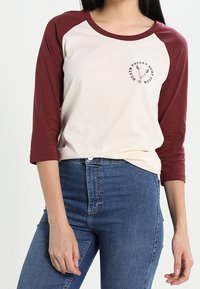 Krämfärgad baseballtröja med vinröda raglanärmar. Har en cirkulär logotyp med ett tjurkranium och text. Tillverkad av mjukt, bomullsmaterial.