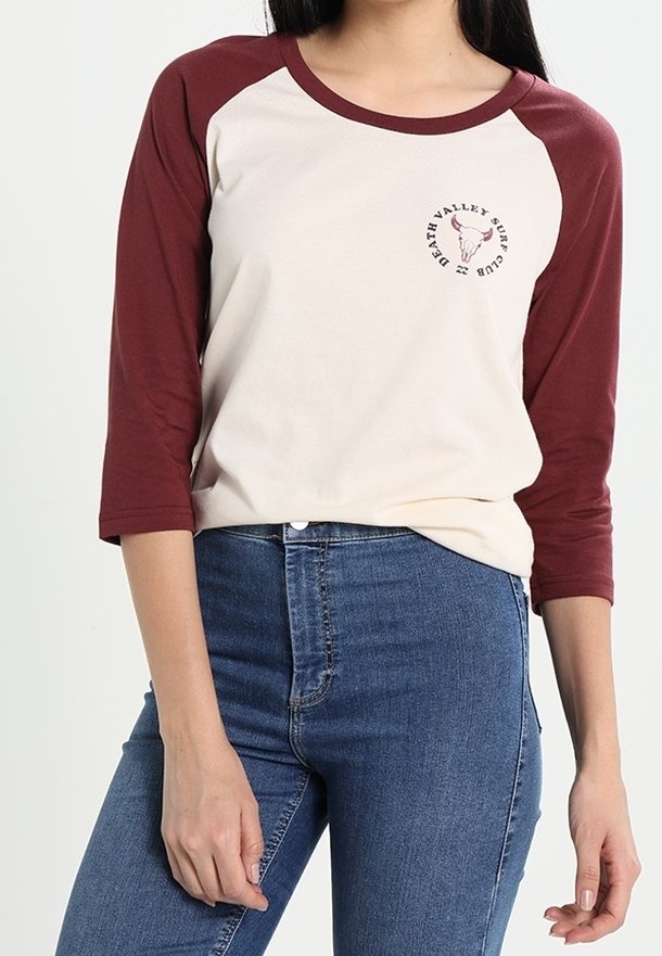 Krämfärgad baseballtröja med vinröda raglanärmar. Har en cirkulär logotyp med ett tjurkranium och text. Tillverkad av mjukt, bomullsmaterial.