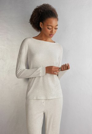 Frau mit lockigem Haar trägt ein hellbeiges, geripptes Langarmshirt und passende Hose, schaut nach unten und richtet ihren Ärmel.