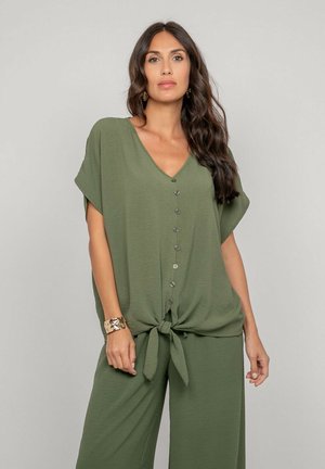 Femme aux longs cheveux bruns portant un haut ample boutonné vert olive noué à la taille et un pantalon large assorti, sur un fond uni.