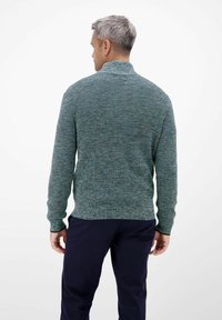 Gestrickter Pullover in gemischten Türkis- und Blautönen, mit hohem Kragen und gerippten Bündchen. Über dunklen Hosen getragen, zeigt die strukturierte Stoffqualität.