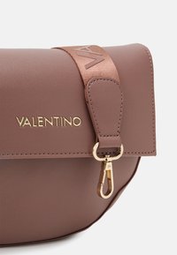 Brun läderväska med ett spänne och rem i guldton. Har präglad logotyp och en slät textur som framhäver en elegant, modern design.