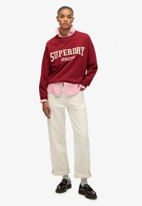Sudadera color burdeos con el texto "SUPERDRY ATHLETIC", superpuesta a una camisa de rayas rosas. Unos pantalones anchos blancos y zapatos oscuros completan el conjunto.