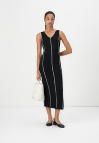 Black sleeveless dress with white vertical stripes, knee-length, made of stretchy fabric. Paired with a light-colored patterned handbag.