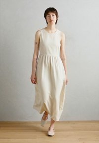 Beige sleeveless dress with a fitted bodice and flowy skirt, made of lightweight fabric. Paired with pale shoes, standing on a wooden floor.