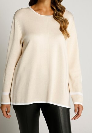 Woman wearing a loose beige long-sleeve sweater with white trim and black leather pants, standing against a plain background.
