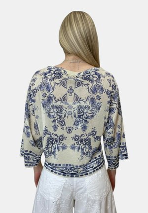 Donna con capelli biondi lisci indossa una maglia beige con motivi floreali blu e pantaloni bianchi ricamati, con la schiena rivolta verso la fotocamera.