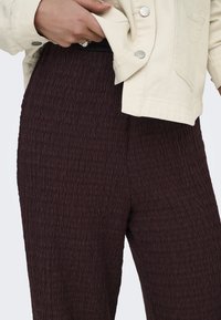 Main avec bague ajustant une veste beige clair portée sur un pantalon marron foncé texturé.