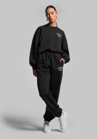 Schwarzes Baumwoll-Sweatshirt und Sweathosen-Set mit lockerer Passform. Mit elastischen Bündchen und einem elastischen Bund, sowie weißen Drucklogo-Details. Weiße Sneakers.