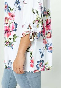 Blouse fleurie en tissu blanc avec des motifs de fleurs roses, bleues et vertes. Les détails noués aux manches complètent une coupe ample et décontractée.