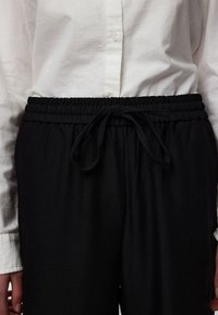 Área de la cintura de una persona que lleva pantalones con cinturilla elástica negra con cordón ajustado y una camisa blanca de manga larga con botones.
