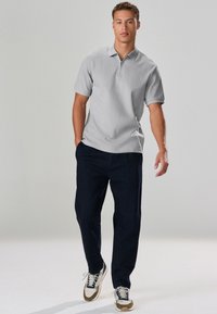 Graues Polohemd mit Reißverschluss, dunkelblaue Hose und graue Sneaker mit hellbraunen Akzenten. Das Model steht vor einem neutralen Hintergrund.