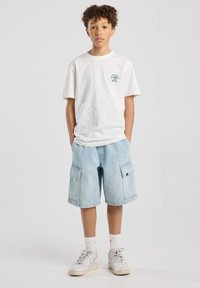 White cotton t-shirt with green graphic, light blue denim cargo shorts with side pockets, and white sneakers with gray accents.