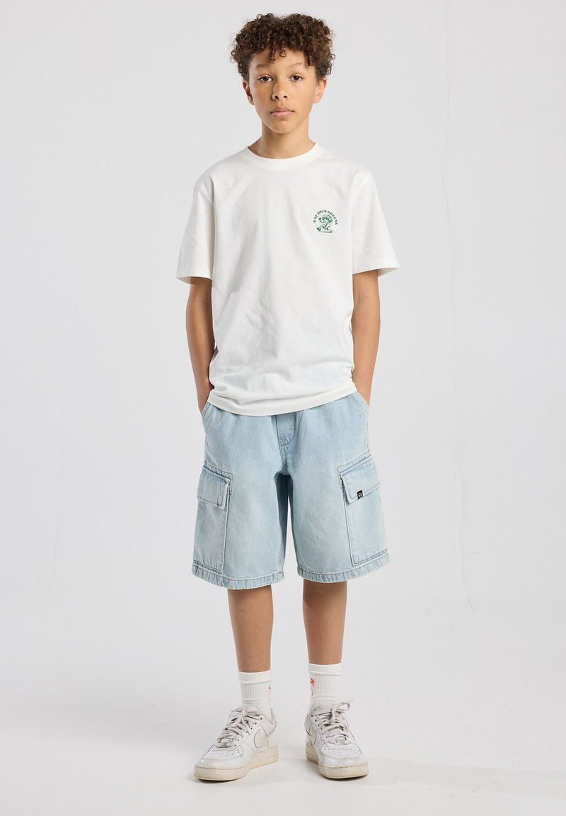 White cotton t-shirt with green graphic, light blue denim cargo shorts with side pockets, and white sneakers with gray accents.