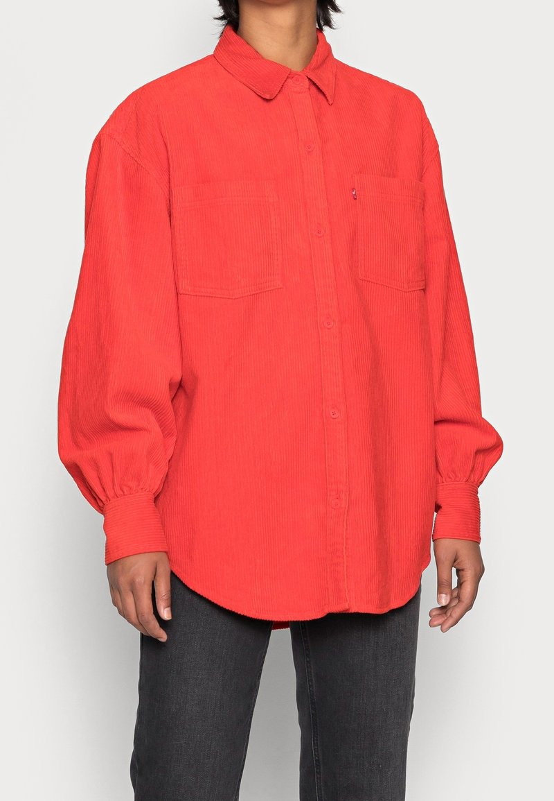 Personne portant une chemise en velours côtelé rouge vif oversize avec fermeture par boutons et deux poches poitrine, portée avec un jean gris foncé.