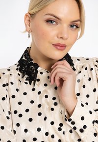 Jonge vrouw met blond haar, gekleed in een crèmekleurige blouse met zwarte stippen en een kanten kraag, die met haar hand haar kin aanraakt.