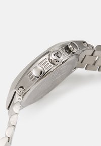 Reloj de acero inoxidable plateado con una esfera texturizada, tres coronas y una correa que presenta eslabones redondeados. Diseño limpio y moderno con acentos minimalistas.