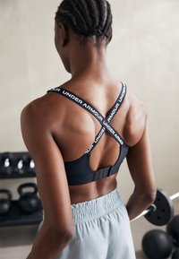 Juodas sportinis liemenukas su kryžminiais petnešomis, puošnias baltu "UNDER ARMOUR" ženklu, saugiu juostelių uždarymu ir lygiu tekstūru.
