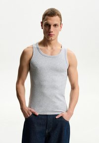 Siv bombažna majica brez rokavov z rebrasto teksturo, okroglo izrezano ovratnikom in širokimi naramnicami; model nosi temne jeans hlače z rokami v žepih.