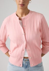 Γυναίκα που φοράει ροζ ζακέτα με πλεκτό σχέδιο πάνω από λευκό cropped τοπ και ανοιχτόχρωμα μπλε τζιν, με τα χέρια πίσω από την πλάτη της.