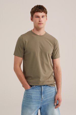 Joven de pie con la mano derecha en el bolsillo, vistiendo una camiseta de manga corta verde oliva y vaqueros azul claro sobre un fondo liso.