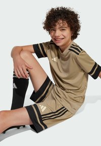 Conjunto de fútbol beige que consiste en una camiseta de manga corta con acentos negros y tres detalles de rayas, acompañado de pantalones cortos y calcetines negros.