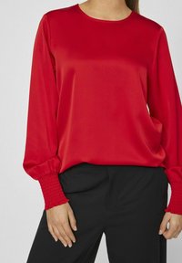 Maglia rossa a maniche lunghe con scollatura rotonda e polsini arricciati, realizzata in un tessuto morbido; abbinata a pantaloni neri.