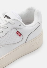 Białe skórzane sneakersy z perforowanymi detalami, płaskimi sznurówkami i czerwonym metkiem Levi's; kremowa gumowa podeszwa z teksturowanymi akcentami.