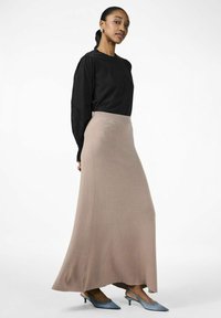 Kvinna klädd i en svart långärmad blus, beige knälång kjol och grå låga slingbacks, stående mot vit bakgrund.