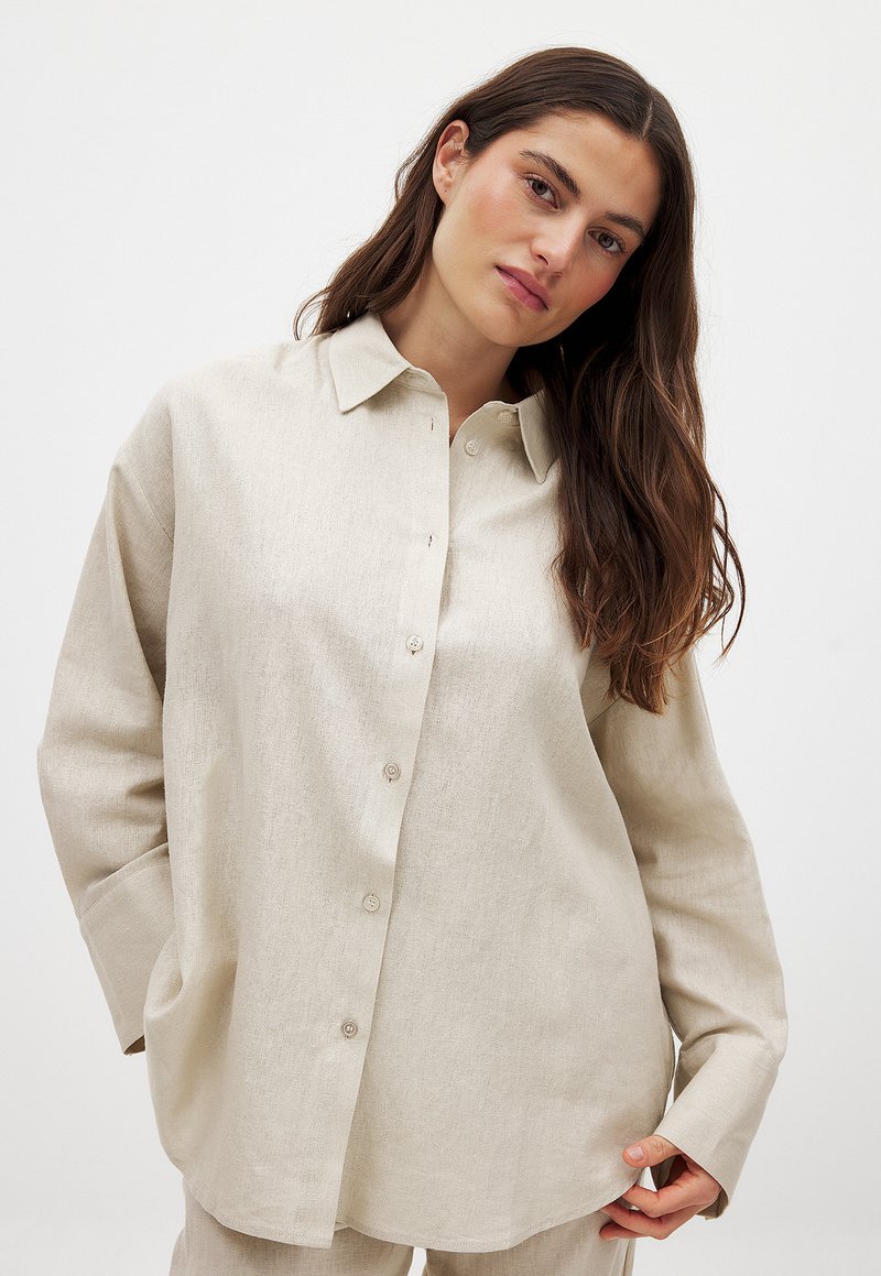 Camicia button-up in lino beige con colletto, maniche lunghe e due tasche frontali. Tessuto morbido con una vestibilità leggermente ampia e orlo arrotondato.