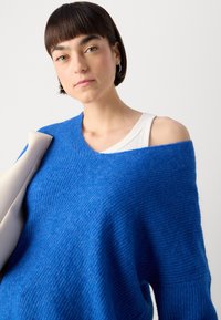 Blue oversized knit sweater with a v-neck and ribbed texture, paired with a white tank top. Model holds a light gray bag.