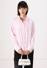 Camicia a maniche lunghe abbottonata color rosa chiaro con sottili righe bianche. Presenta un colletto a punta e una vestibilità oversize. Abbinata a una piccola borsa bianca.