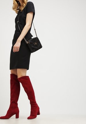 Stivali over-the-knee in suede rosso con punta leggermente appuntita e tacco spesso. Abbinati a un vestito nero aderente e una piccola borsa nera.