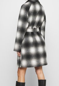 Cappotto a quadri bianco e nero con vita incinturata, maniche lunghe e tessuto strutturato. Presenta un colletto e una silhouette leggermente svasata.