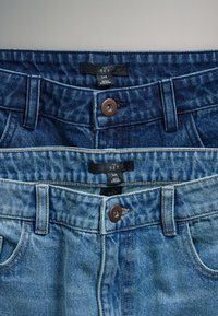 Twee paar blauwe denimjeans gestapeld, met taillebanden met knoopsluitingen en zwarte maatlabels met de tekst "The Set 34R."