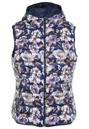 Person iført en grå langærmet skjorte, en marineblå blomsterquiltet vest med hætte, hvide skibukser og sorte skistøvler med røde såler.