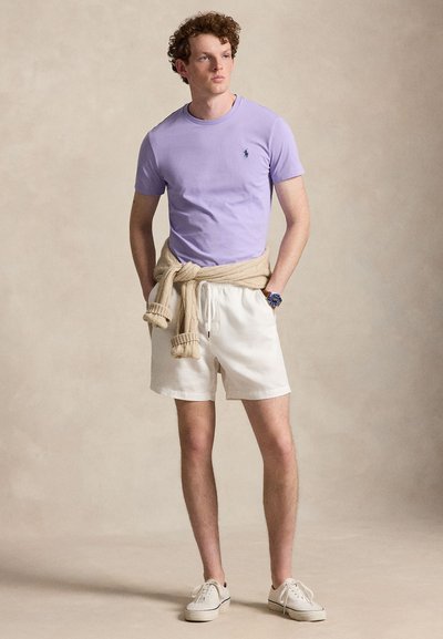 Fiatal férfi zsebre tett kézzel áll, lila pólót, bézs rövidnadrágot, a derekán kötött krémszínű pulóvert, fehér tornacipőt és kék órát visel.