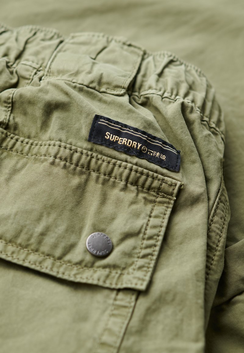 Olijfgroen stof met elastische tailleband, zakflap beveiligd met een gemerkte metalen drukknoop en een zwart label met de tekst "SUPERDRY" in gouden letters.