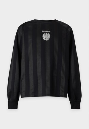 Mand stående iført sort langærmet stribet skjorte med hvidt emblem, løse sorte bukser og sorte sneakers på hvid baggrund.