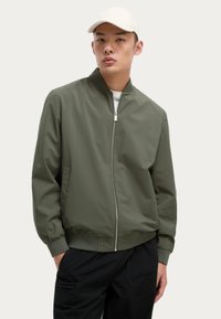 Chaqueta bomber verde oliva con cierre frontal, cuello y puños acanalados, con bolsillos laterales, llevada sobre una camisa blanca y pantalones negros.