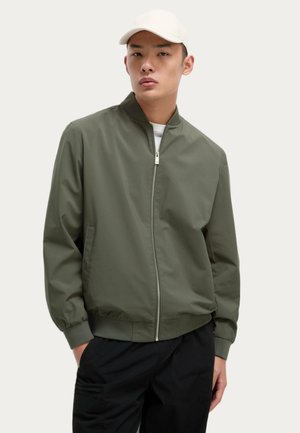 Chaqueta bomber verde oliva con cierre frontal, cuello y puños acanalados, con bolsillos laterales, llevada sobre una camisa blanca y pantalones negros.