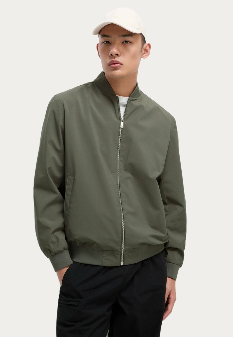 Chaqueta bomber verde oliva con cierre frontal, cuello y puños acanalados, con bolsillos laterales, llevada sobre una camisa blanca y pantalones negros.