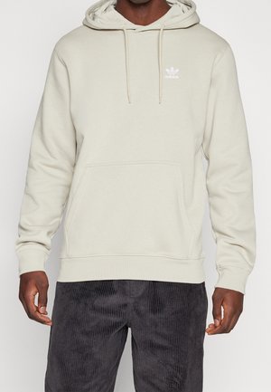 Mężczyzna w jasnobeżowej bluzie Adidas z kapturem i przednią kieszenią oraz czarnych spodniach sztruksowych, widoczny od ramion do połowy uda.