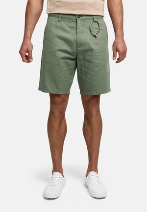 Homme portant un short chino vert olive avec un porte-clés tressé, un haut beige et des baskets blanches, debout devant un fond uni.