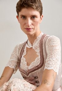 Koronkowa bluzka z falbaniastym dekoltem, z różową warstwą gorsetu, ozdobiona wstążkowymi akcentami i dekoracyjnymi guzikami. Widoczna szczegółowa haftowana florystyka.