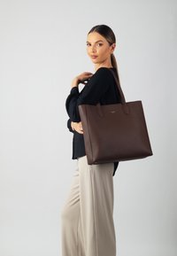 Brown textured leather tote bag with two sturdy handles, subtle logo, and a structured shape, showcased against a neutral background.
