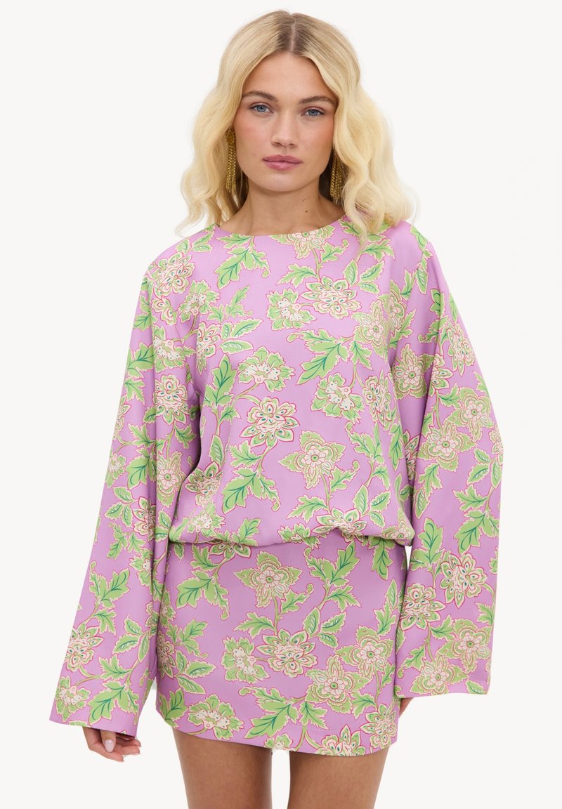 Femme blonde portant une robe longue à manches longues rose à motifs floraux avec des feuilles vertes et des fleurs beige, debout sur un fond blanc uni.