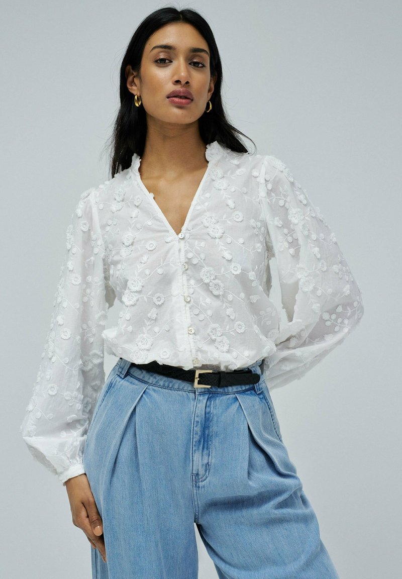 Blusa blanca con bordado floral texturizado, escote en V, mangas abullonadas y cierre de botones, combinada con pantalones de mezclilla de cintura alta.
