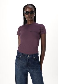 Lilla tætsiddende T-shirt med korte ærmer og et lille logo på brystet, parret med højttaljede mørkeblå jeans.