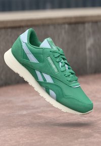 Zaļas kedas ar sintētisku augšdaļu, zamša akcentiem, baltām svītrām un plakanu šņorējumu. Gumijas zole ar redzamu rakstu un Reebok zīmolu.